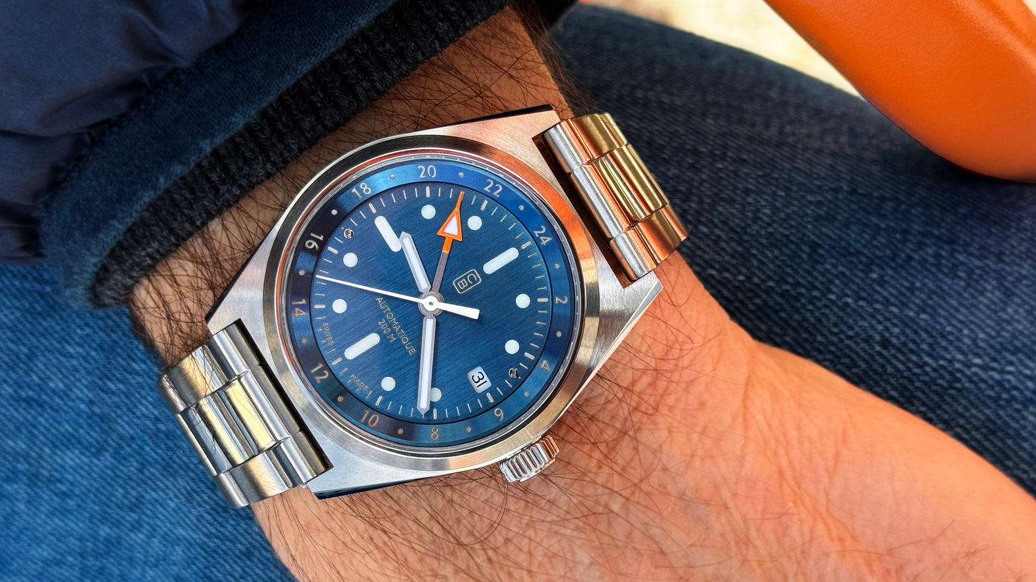 A full recycled stainless steel wristwatch with a blue dial on repurposed stainless steel bracelet, worn on the wrist. Blue jeans denim as background